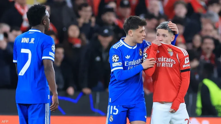 Two Real Madrid players in blue jerseys comfort a teammate in a red Benfica jersey on the field while a crowd watches.
