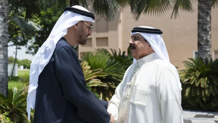 Two men in traditional dress shake hands outdoors, facing each other in front of palm trees and a beige building.