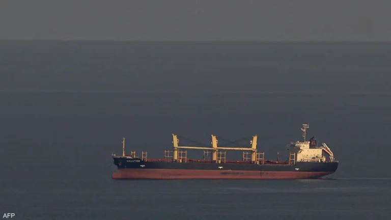 Large cargo ship with yellow cranes gliding on calm open water, red hull and white superstructure at the rear.
